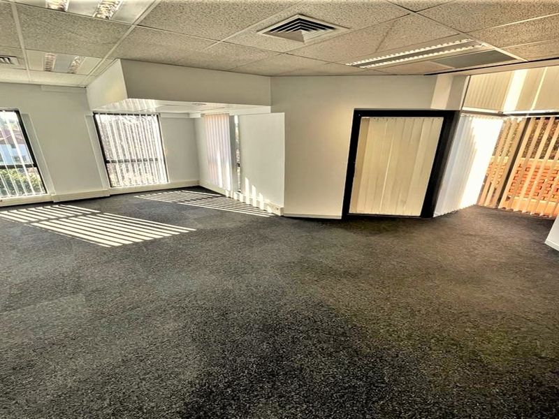 A spacious, empty office area featuring carpeted flooring, suspended ceilings with integrated lighting, and large windows with vertical blinds providing natural light.