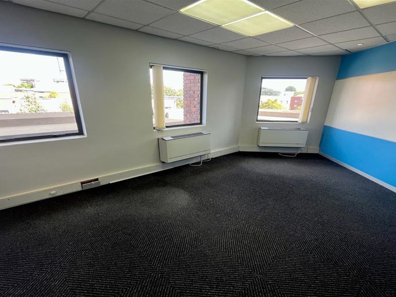 A vacant office space featuring carpeted flooring, suspended ceiling with recessed lighting, and wall-mounted HVAC units with natural light from multiple windows.