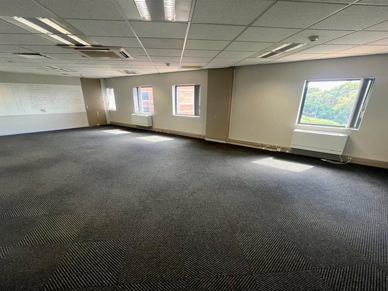 A spacious, empty office area featuring carpeted flooring, suspended ceilings with recessed lighting, and wall-mounted HVAC units.