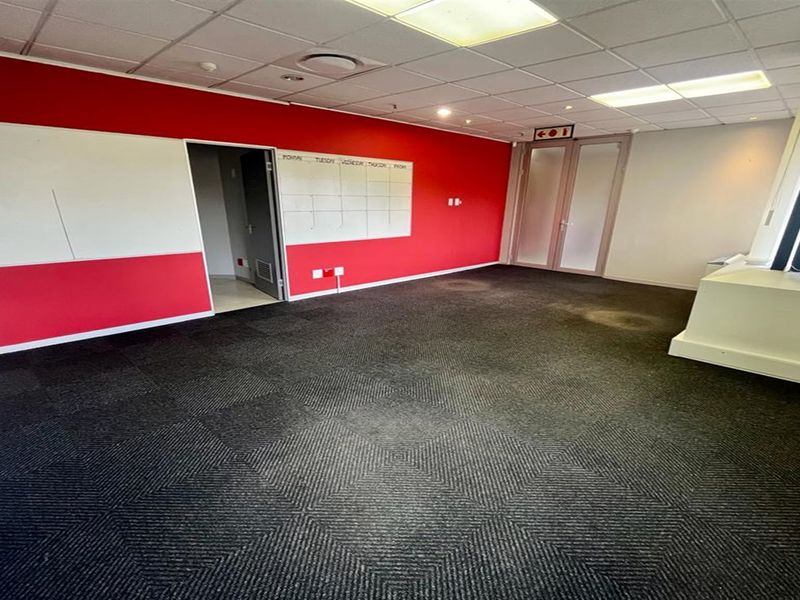An empty office space featuring dark carpet tiles, a suspended ceiling with recessed lighting, and a prominent red accent wall with a built-in whiteboard.