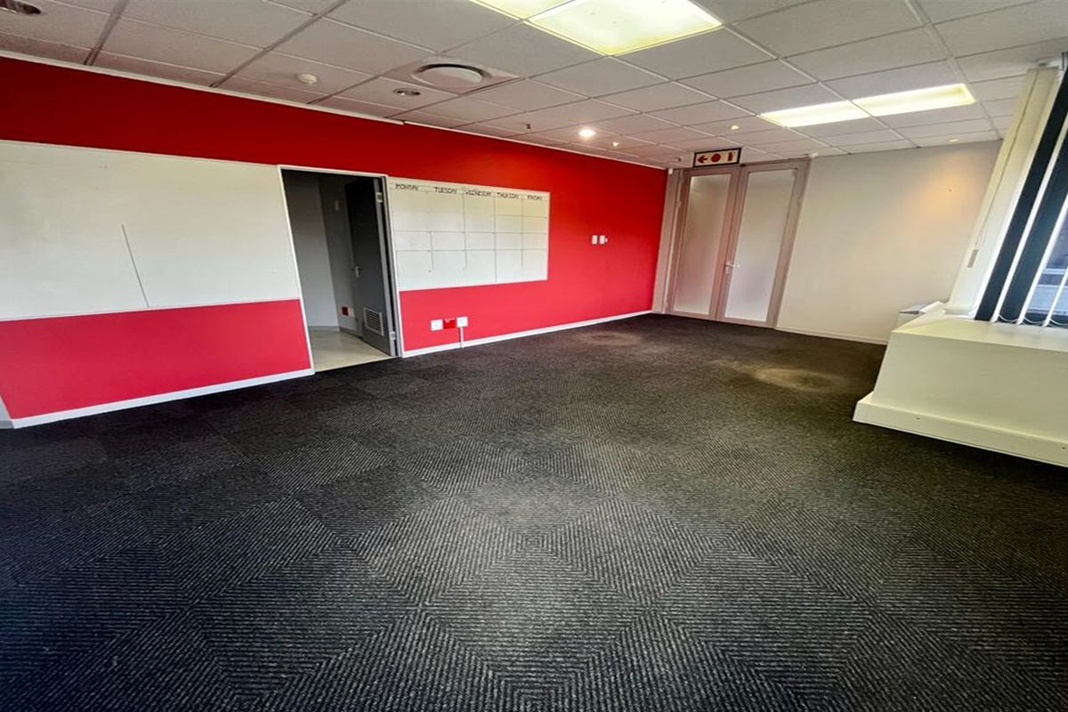 An empty office space featuring dark carpet tiles, a suspended ceiling with recessed lighting, and a prominent red accent wall with a built-in whiteboard.