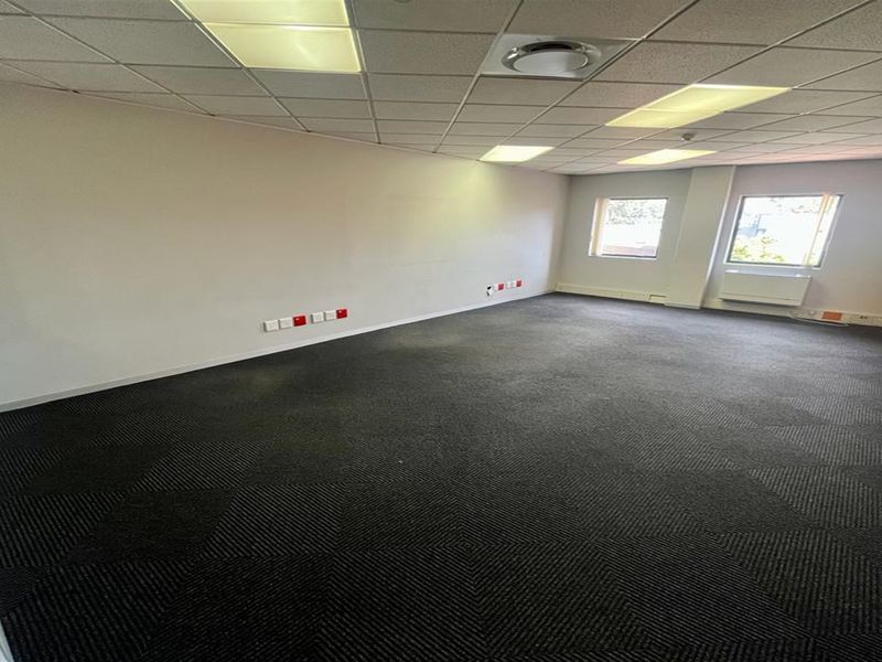 A spacious, empty office area featuring carpeted flooring, a suspended ceiling with recessed lighting, and integrated wall-mounted data and power points.