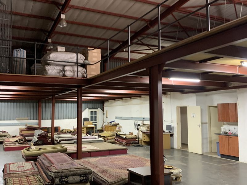 Spacious industrial warehouse area featuring a steel mezzanine level, exposed roof trusses, concrete flooring, and a functional kitchenette corner.