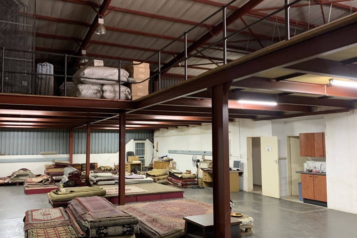 Spacious industrial warehouse area featuring a steel mezzanine level, exposed roof trusses, concrete flooring, and a functional kitchenette corner.