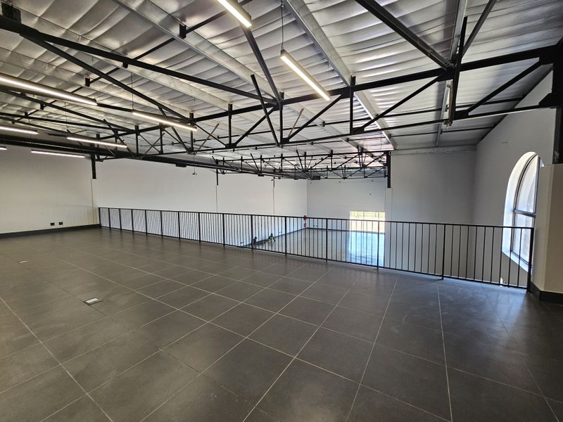 Spacious open-plan mezzanine level featuring tiled flooring, exposed industrial steel roof trusses, and modern linear pendant lighting overlooking the lower retail area.