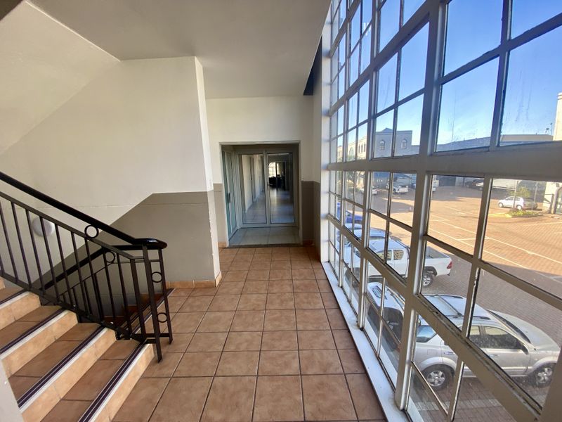 A bright, tiled hallway featuring a metal staircase and expansive steel-framed windows overlooking the exterior parking area.