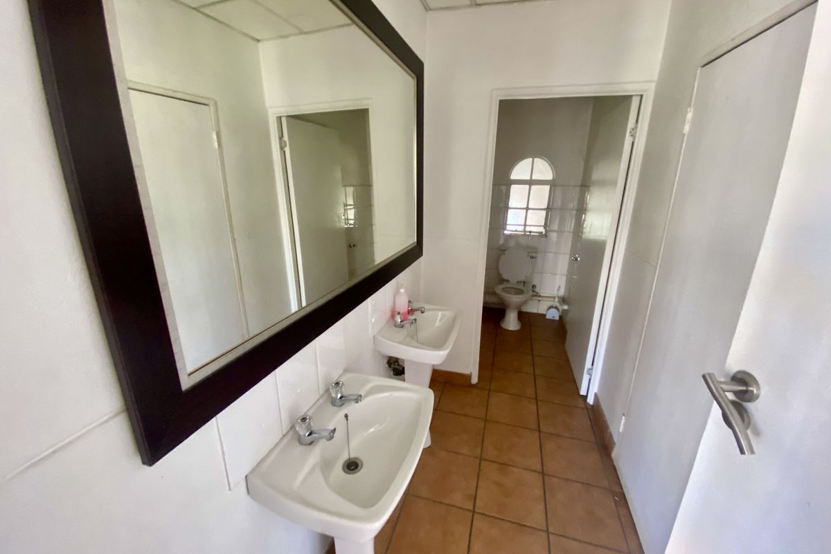 A functional industrial-style bathroom featuring dual pedestal wash basins, a large wall mirror, and a separate toilet area with tiled flooring.