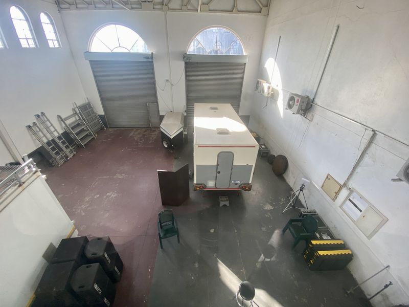Spacious industrial garage featuring high ceilings with exposed trusses, dual roller shutter doors, arched windows, and wall-mounted air conditioning units.