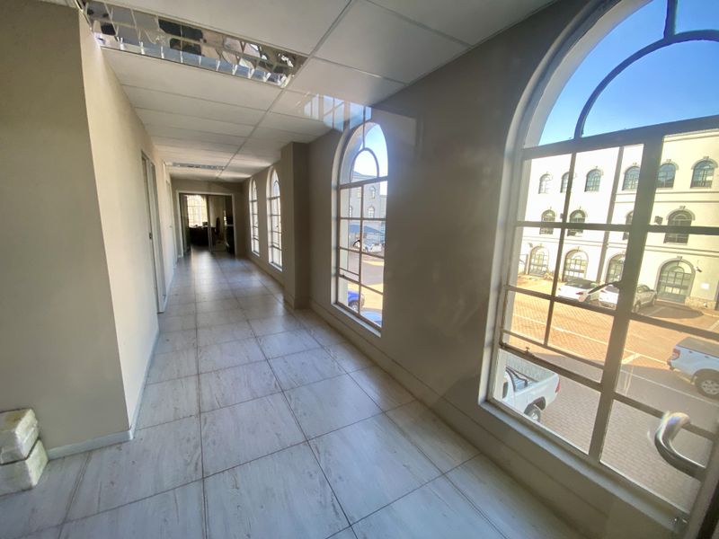A spacious, empty industrial hallway featuring tiled flooring, a suspended ceiling with recessed lighting, and large arched windows overlooking the exterior parking area.