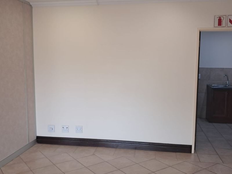 A clean, empty office space featuring tiled flooring, neutral walls, and a doorway leading to a small kitchenette area with a sink and cabinetry.
