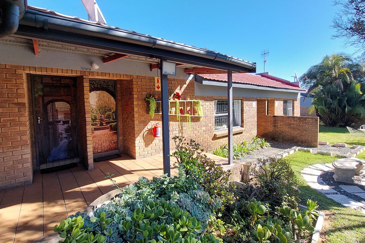 A rear exterior view of the guest house featuring a brick facade, covered patio area, and a landscaped garden with a paved pathway.