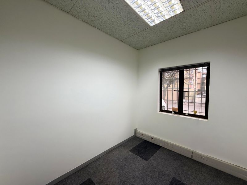 A vacant office space featuring carpet flooring, a suspended ceiling with recessed lighting, and a window fitted with security bars.