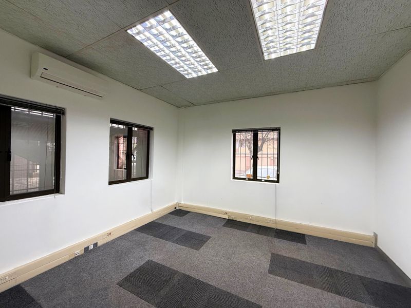 A clean, empty office space featuring carpet tiles, recessed ceiling lighting, split-system air conditioning, and windows with security bars.