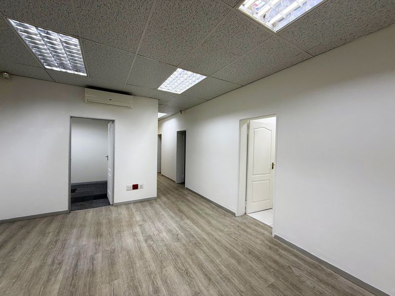 A clean, vacant office space featuring modern laminate flooring, a suspended ceiling with recessed lighting, and a wall-mounted air conditioning unit.