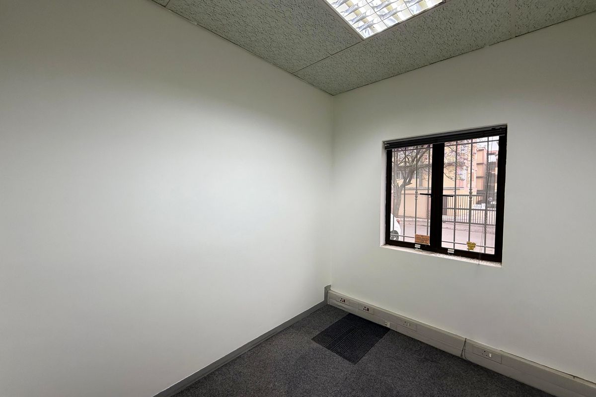 A vacant office space featuring carpet flooring, a suspended ceiling with recessed lighting, and a window fitted with security bars.