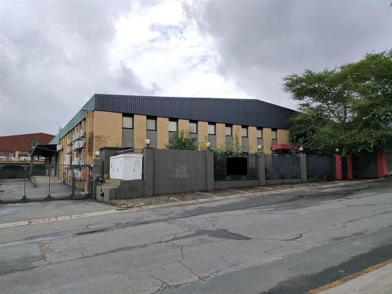Front exterior view of an industrial warehouse building featuring a brick facade, metal roofing, perimeter security wall, and gated access.