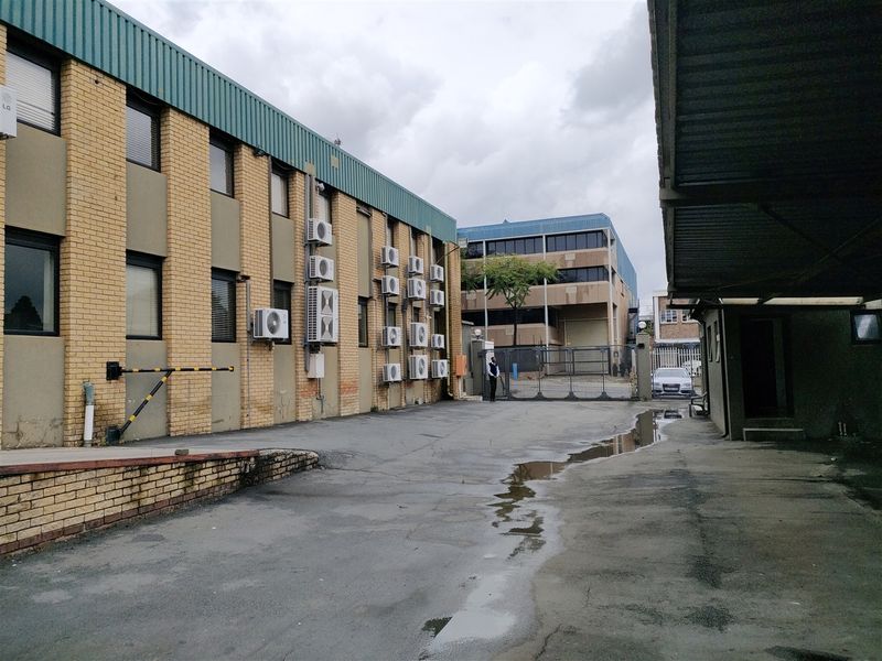 Exterior view of an industrial property featuring a brick facade, multiple wall-mounted air conditioning units, and a paved driveway area with security access gate.