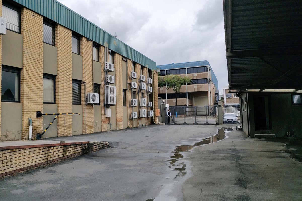 Exterior view of an industrial property featuring a brick facade, multiple wall-mounted air conditioning units, and a paved driveway area with security access gate.
