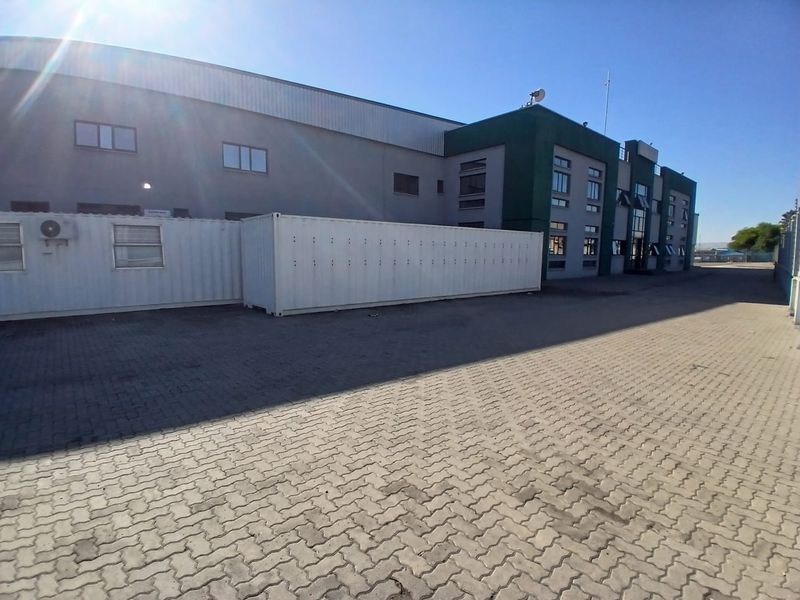 Exterior view of an industrial facility featuring a paved yard, container storage units, and a multi-story office and warehouse building structure.