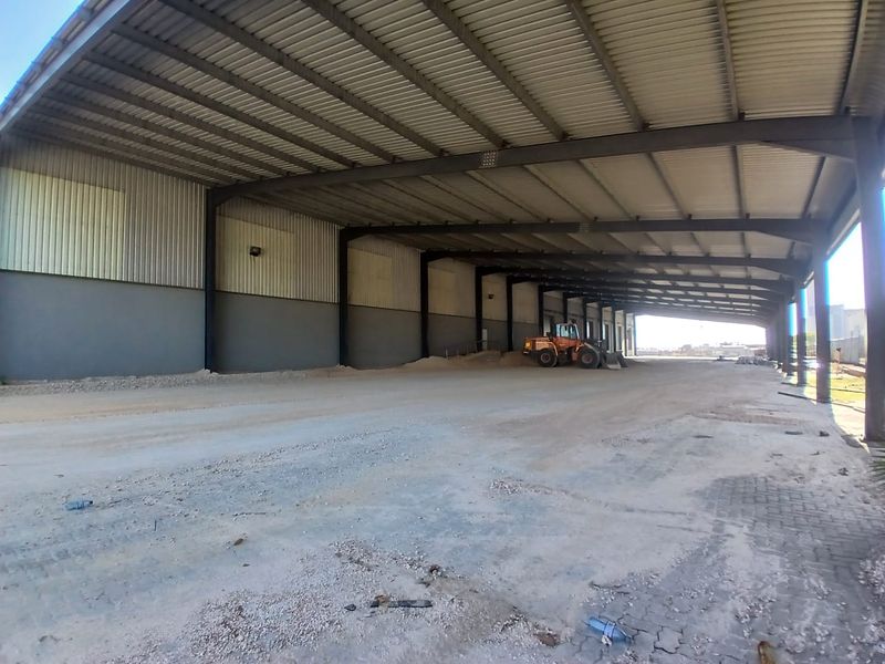 Large industrial covered outdoor storage or loading area featuring a high steel-frame roof structure and expansive concrete and gravel flooring.