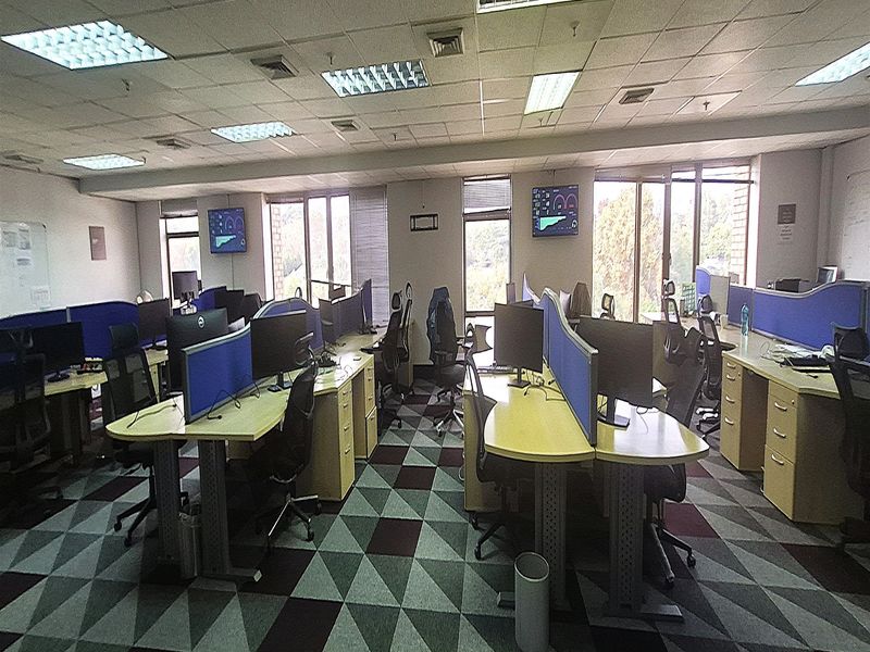 A spacious open-plan office area featuring suspended ceilings, recessed lighting, large windows providing natural light, and patterned carpet flooring.
