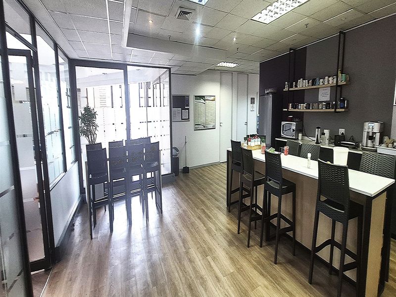 Modern office breakroom featuring a built-in kitchenette counter, laminate flooring, glass partitions, and recessed lighting in a professional workspace setting.