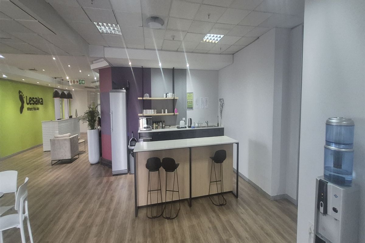 Modern office space featuring a functional kitchenette area with built-in shelving, laminate flooring, and a suspended ceiling with recessed lighting.