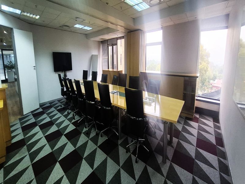 A professional office boardroom featuring a large central table, built-in storage cabinetry, patterned carpet flooring, and ample natural light from large windows.