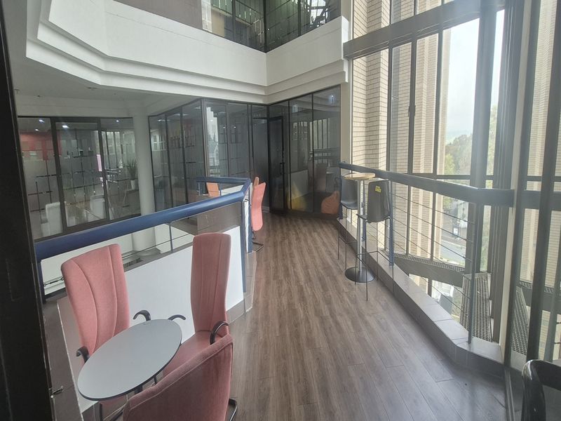 Modern office mezzanine area featuring full-height glass windows, laminate flooring, and glass partitions overlooking a multi-level atrium space.