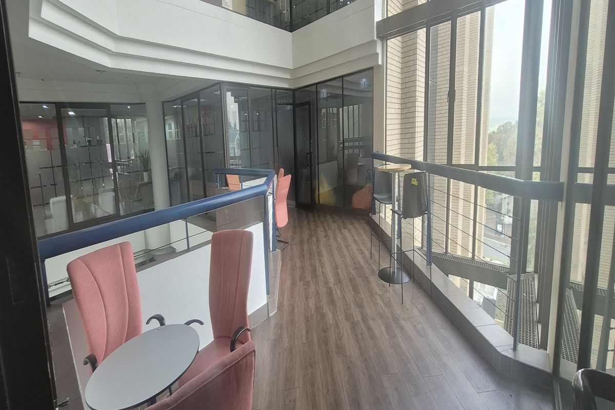 Modern office mezzanine area featuring full-height glass windows, laminate flooring, and glass partitions overlooking a multi-level atrium space.