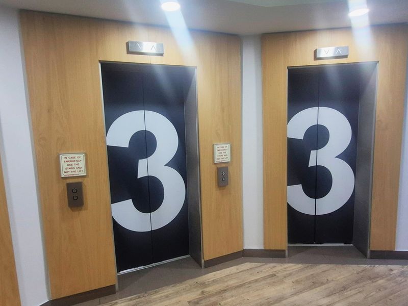 A clean and modern office building elevator lobby featuring dual elevators with wood veneer paneling and clear floor level signage.