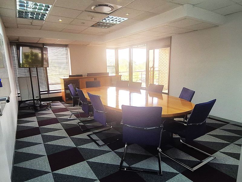 A professional office boardroom featuring a large central table, patterned carpet tiles, and ample natural light from large windows with venetian blinds.