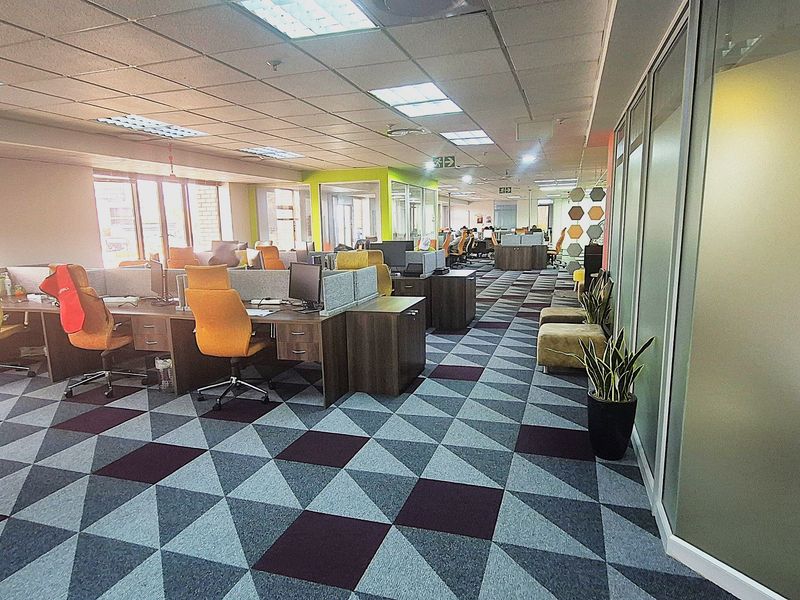 A spacious open-plan office area featuring modern glass partitions, suspended ceilings with recessed lighting, and durable patterned carpet tiles throughout the workspace.