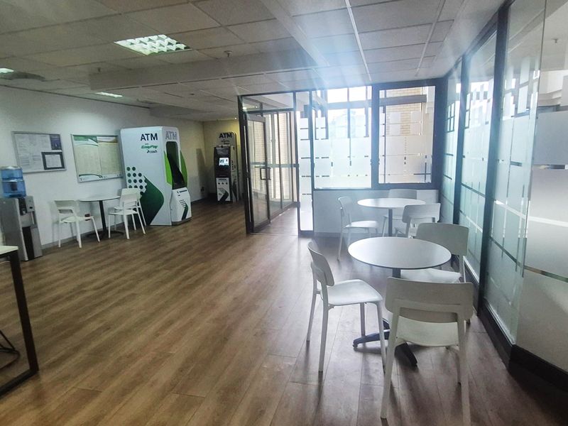 Modern office lobby or waiting area featuring glass partitions, suspended ceilings, and wood-look laminate flooring with integrated ATM facilities.
