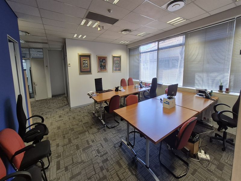 A functional office space featuring carpeted flooring, suspended ceiling with recessed lighting, and large windows with venetian blinds for natural light.