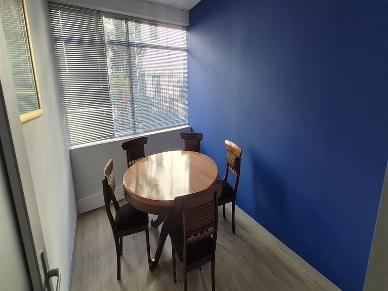 A compact office space featuring light wood-look laminate flooring, a bold blue accent wall, and large windows fitted with venetian blinds.