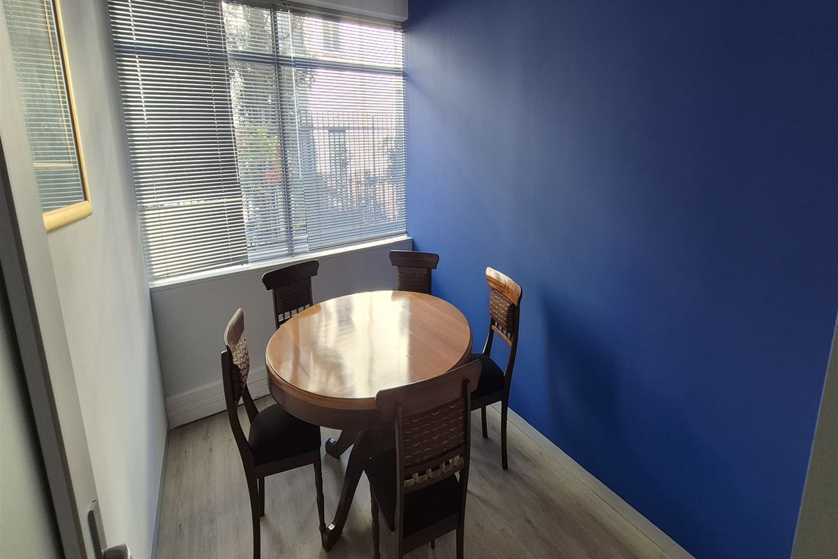 A compact office space featuring light wood-look laminate flooring, a bold blue accent wall, and large windows fitted with venetian blinds.