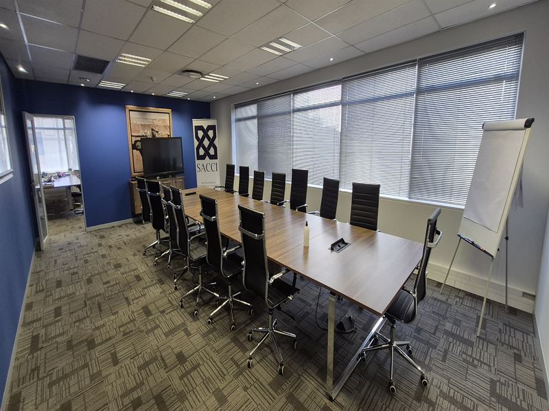A professional boardroom featuring a large conference table, suspended ceiling with recessed lighting, carpet tiles, and large windows with venetian blinds.