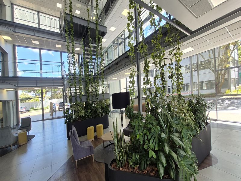 Modern office building entrance lobby featuring a double-volume atrium, steel staircase, extensive glass facade, and contemporary tiled flooring.
