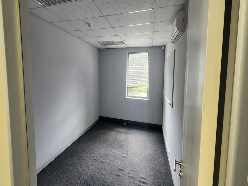 A compact, empty office space featuring a suspended ceiling, recessed lighting, wall-mounted air conditioning, and carpeted flooring.