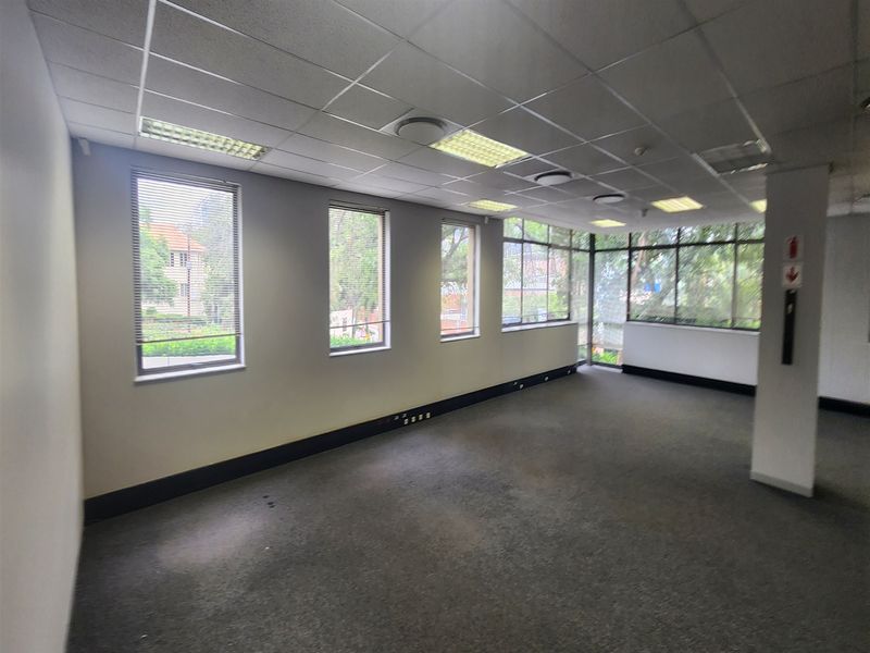 An empty, open-plan office space featuring large windows, suspended ceilings with recessed lighting, and carpeted flooring.