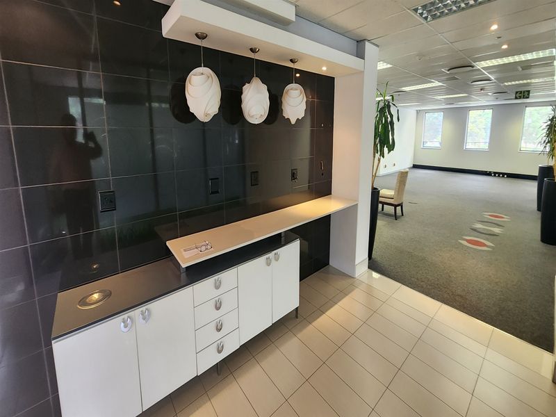 A professional office space featuring a built-in kitchenette or refreshment station with modern cabinetry, tiled flooring, and a suspended ceiling with recessed lighting.