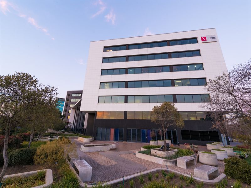 A modern multi-story office building facade featuring large tinted windows, set within a well-maintained landscaped garden with concrete planters and paved walkways.