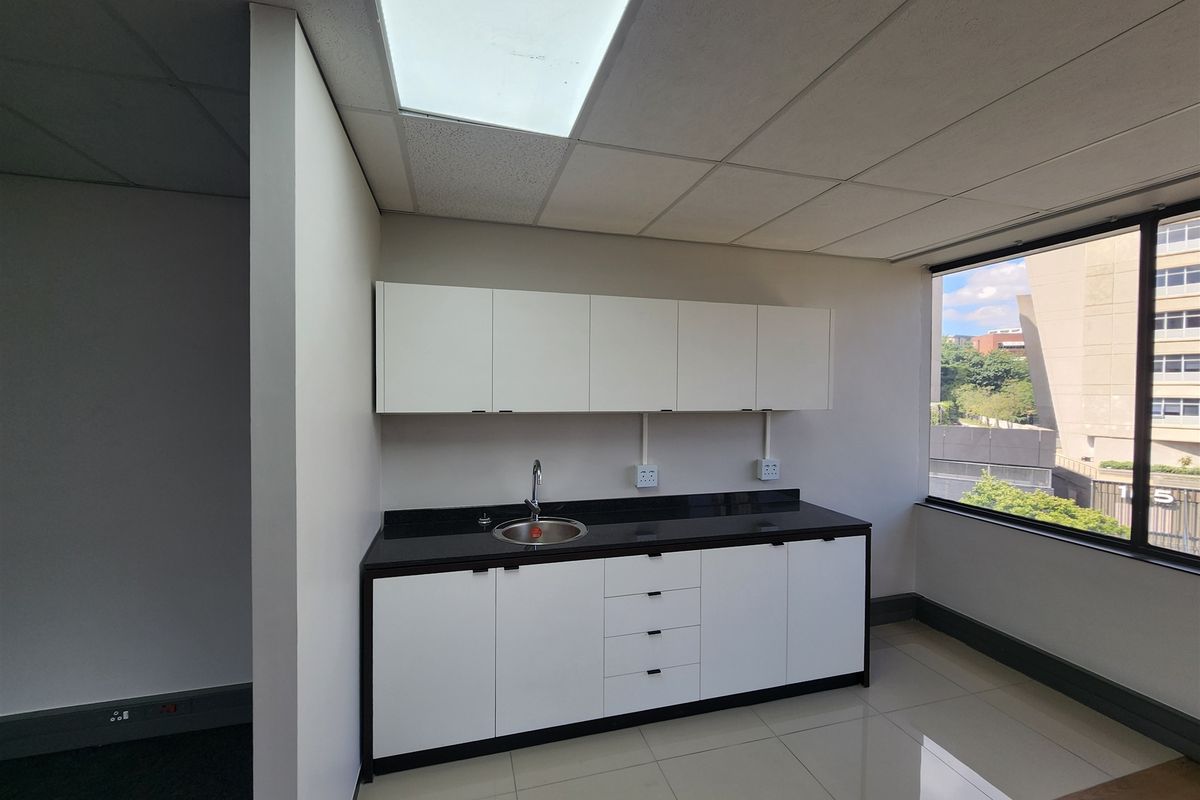 A clean, modern office kitchenette featuring white cabinetry, black granite countertops, a stainless steel sink, and large windows providing natural light.