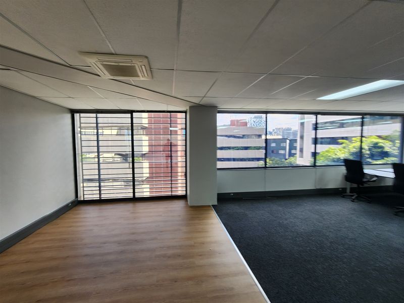 A bright, open-plan office space featuring a mix of carpet and laminate flooring, large windows with security bars, and a suspended ceiling.