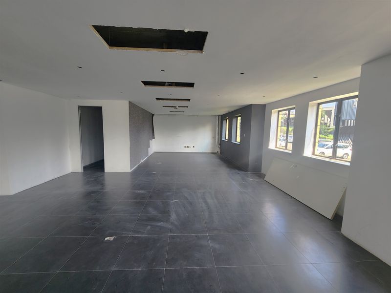 An empty, open-plan office space featuring dark tiled flooring, large windows, and an unfinished ceiling with exposed cutouts.