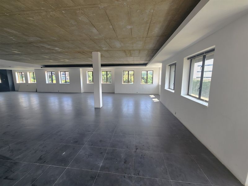 A spacious, open-plan office area featuring an exposed concrete ceiling, large format floor tiles, and abundant natural light from multiple windows.