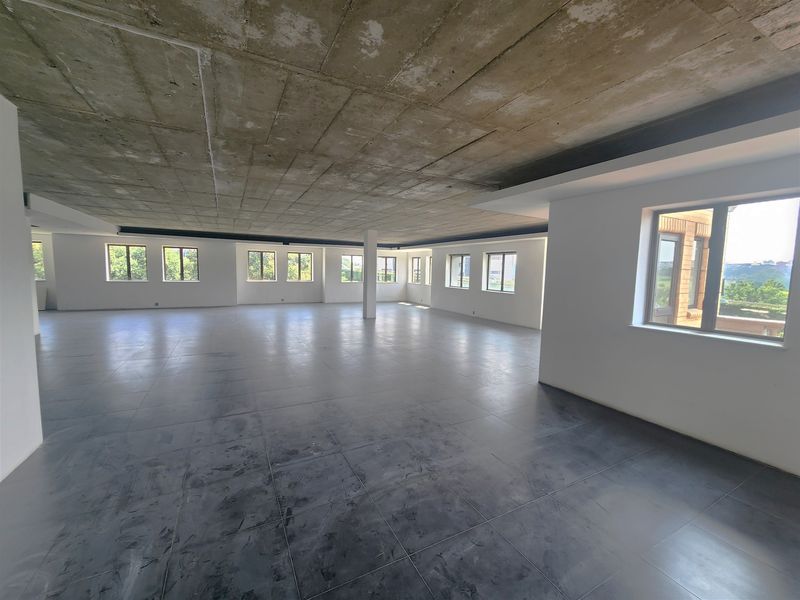 A spacious, open-plan office area featuring an exposed concrete ceiling, large format floor tiles, and abundant natural light from multiple windows.
