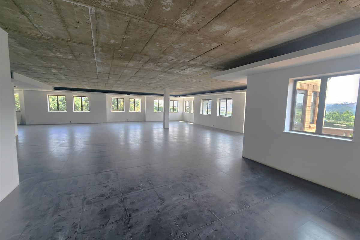 A spacious, open-plan office area featuring an exposed concrete ceiling, large format floor tiles, and abundant natural light from multiple windows.