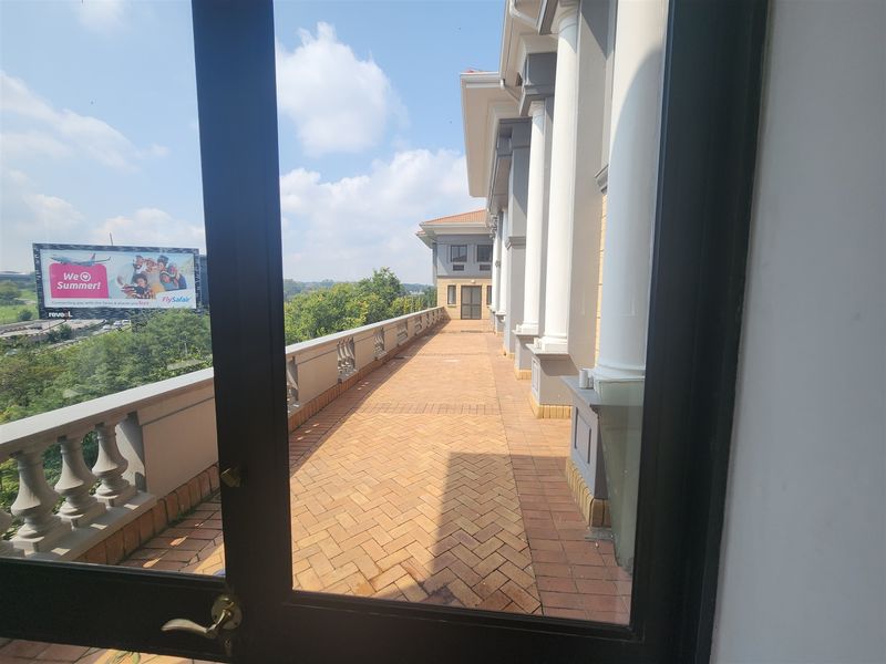 A spacious outdoor balcony featuring herringbone brick paving, classical columns, and a balustrade, offering an elevated view of the surrounding area.